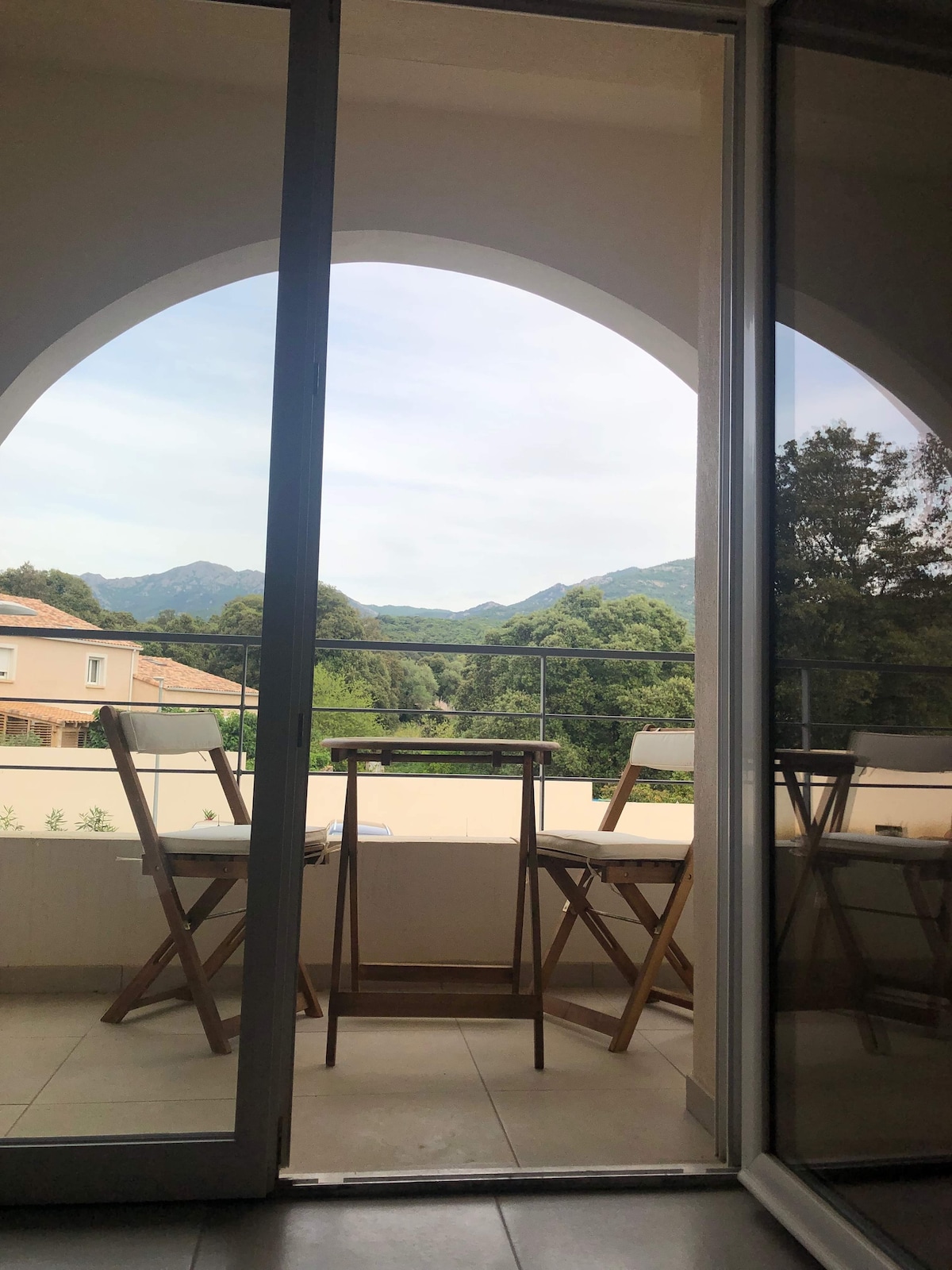 A terrace with a wooden table and two folding chairs can be seen, framed by a large arched window. The view includes lush green mountains in the background, offering a serene natural landscape. The surrounding area features building exteriors with soft colors.