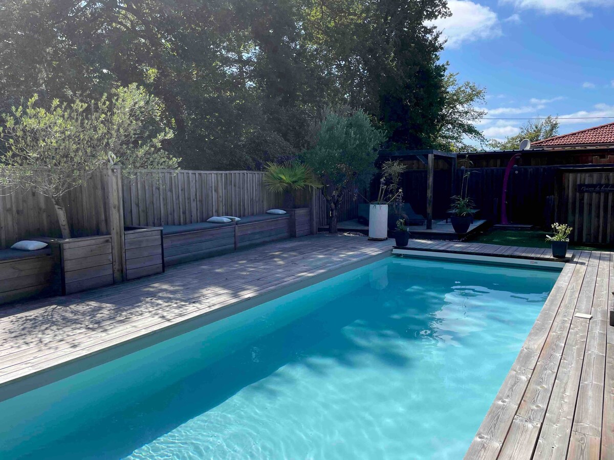 A tranquil pool area is surrounded by wooden decking, offering a refreshing blue water feature. Green plants and potted greenery are positioned along the perimeter, enhancing the natural setting. An outdoor shower is visible in the background, adding to the convenience of this private retreat.
