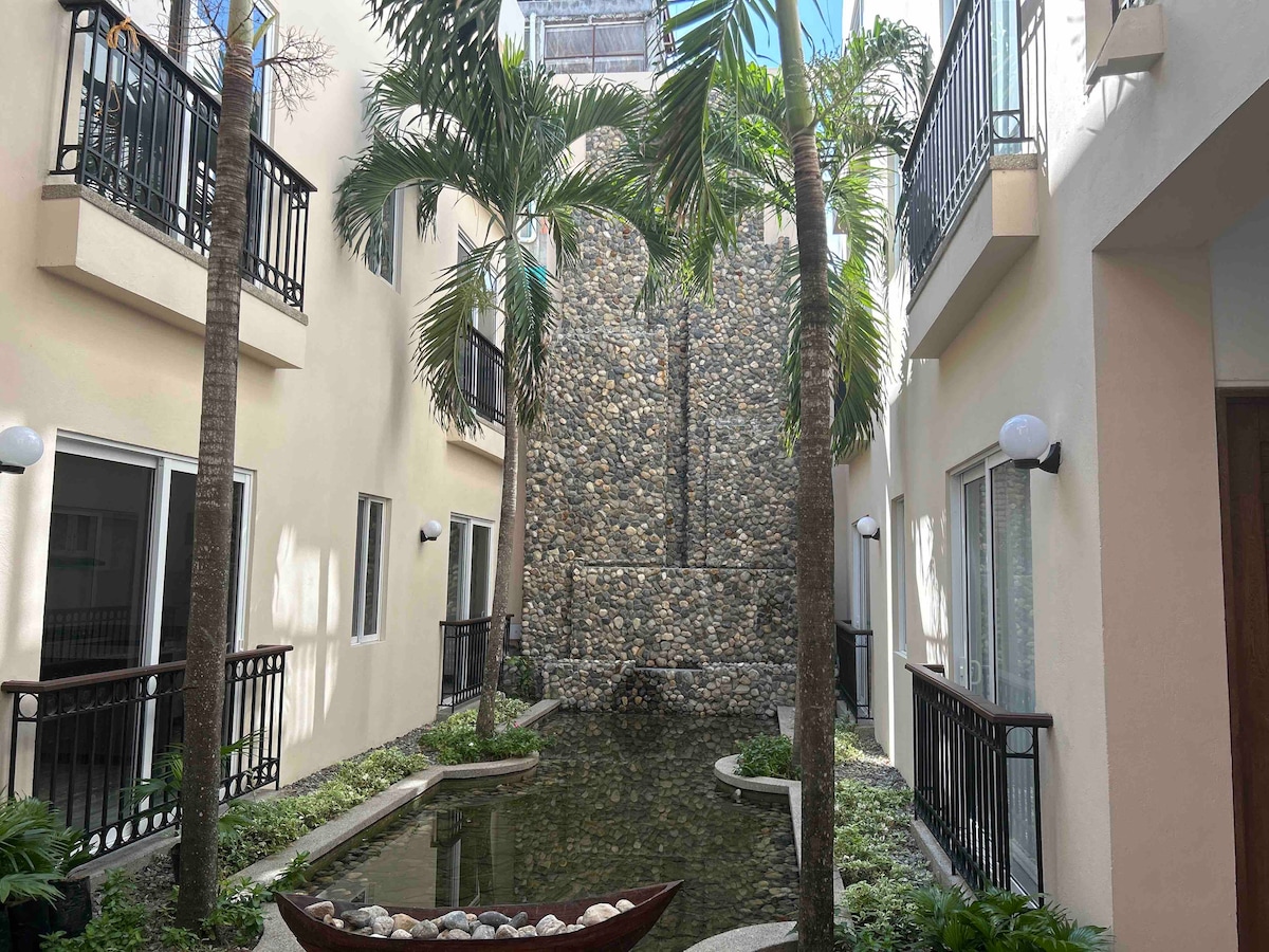 A serene courtyard features a stone waterfall surrounded by lush greenery and palm trees. The clear water reflects the structure's neutral walls and is bordered by smooth stones. Balconies can be seen overlooking the calm setting, enhancing the tranquil atmosphere.