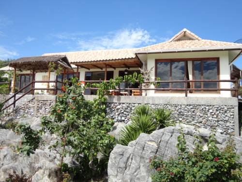 The villa features a stone foundation with a wooden deck, surrounded by lush tropical vegetation. Large windows let in natural light, and a thatched roof adds to the island charm. A rocky landscape complements the structure, providing a natural backdrop.