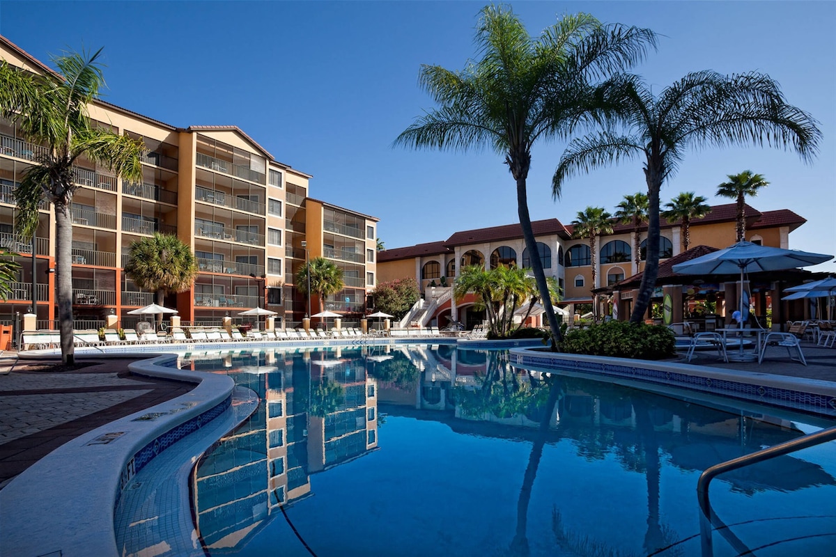 A large outdoor swimming pool is surrounded by lounge chairs and umbrellas. Palm trees frame the scene, highlighting the resort's architecture in the background. The clear blue water reflects the bright sky, creating a relaxing atmosphere in a well-maintained area.