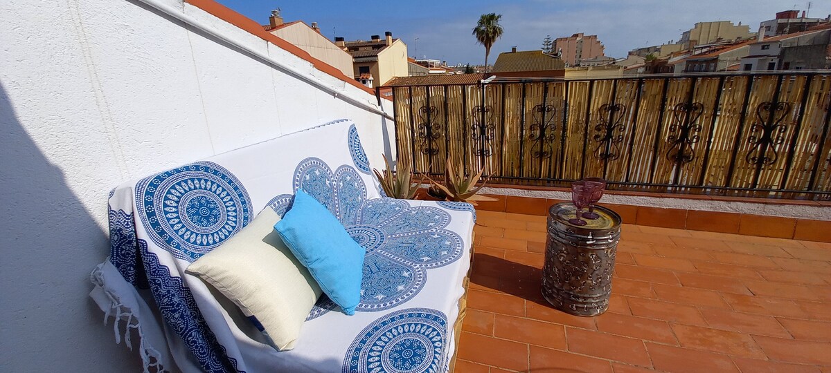 A sunlit terrace features a comfortable seating area with a decorative blanket and cushions. Colorful potted plants are positioned nearby, and a small table adds to the inviting space, offering views of the surrounding neighborhood and palm trees.