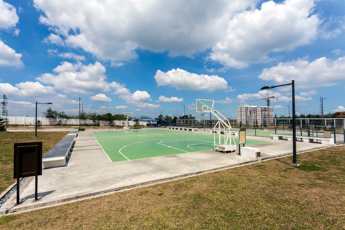 A spacious outdoor basketball court is shown, featuring a bright green playing surface. Two basketball hoops are positioned at opposite ends, surrounded by well-maintained grass and a clear blue sky with scattered clouds. Seating is available along the perimeter.
