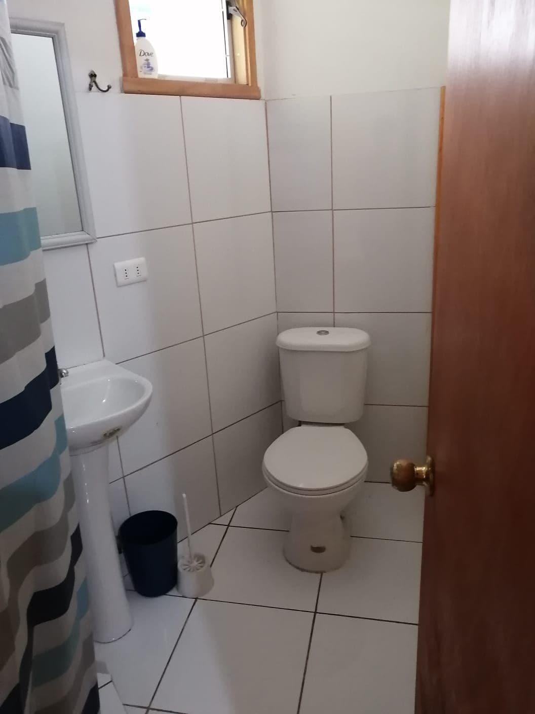 A compact bathroom is featured, with white tiled walls and floor. A sink with a mirror is positioned beside a toilet. A striped shower curtain adds a touch of color, while essential toiletries are visible on the sink. Natural light enters through a small window.