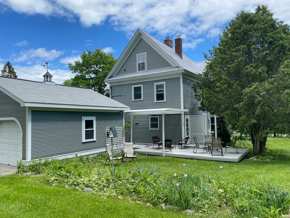 A two-story gray house stands beside a well-maintained lawn, featuring a spacious back deck with seating and a pergola. Flower beds line the area, enhancing the outdoor space, while a garage is visible to the left, framed by greenery under a partially cloudy sky.