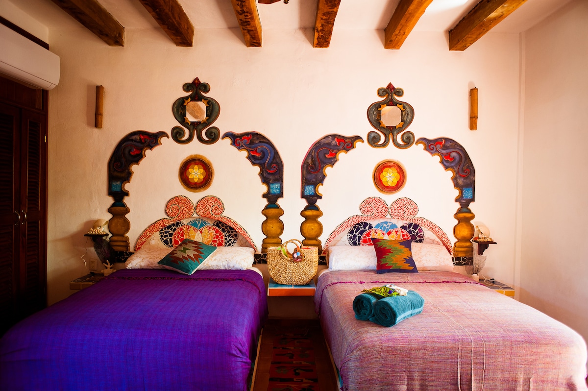 The cozy sleeping area features two beds adorned with colorful blankets. Unique wall art with intricate designs highlights the space, complemented by wooden beams above. Soft towels are neatly arranged on the beds alongside decorative pillows, creating a welcoming environment.