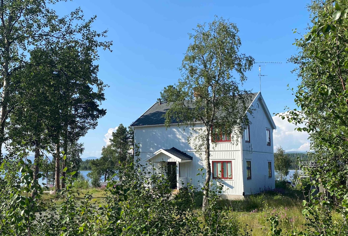 The white house is surrounded by greenery, with a clear blue sky above. The structure features multiple windows, creating a contrast with the natural landscape. Nearby, trees and bushes frame the entrance, providing a sense of privacy and connection to the outdoors.