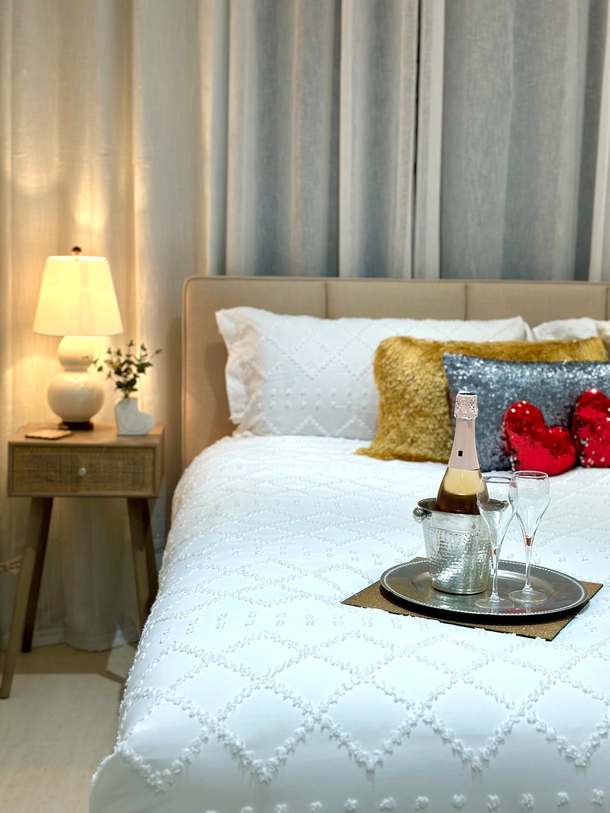A cozy bedroom features a spacious bed with a textured white comforter. A bedside table displays a lamp and a small plant. On the bed, a metal tray holds a champagne bottle, two glasses, and ice in a bucket, adding a touch of elegance.