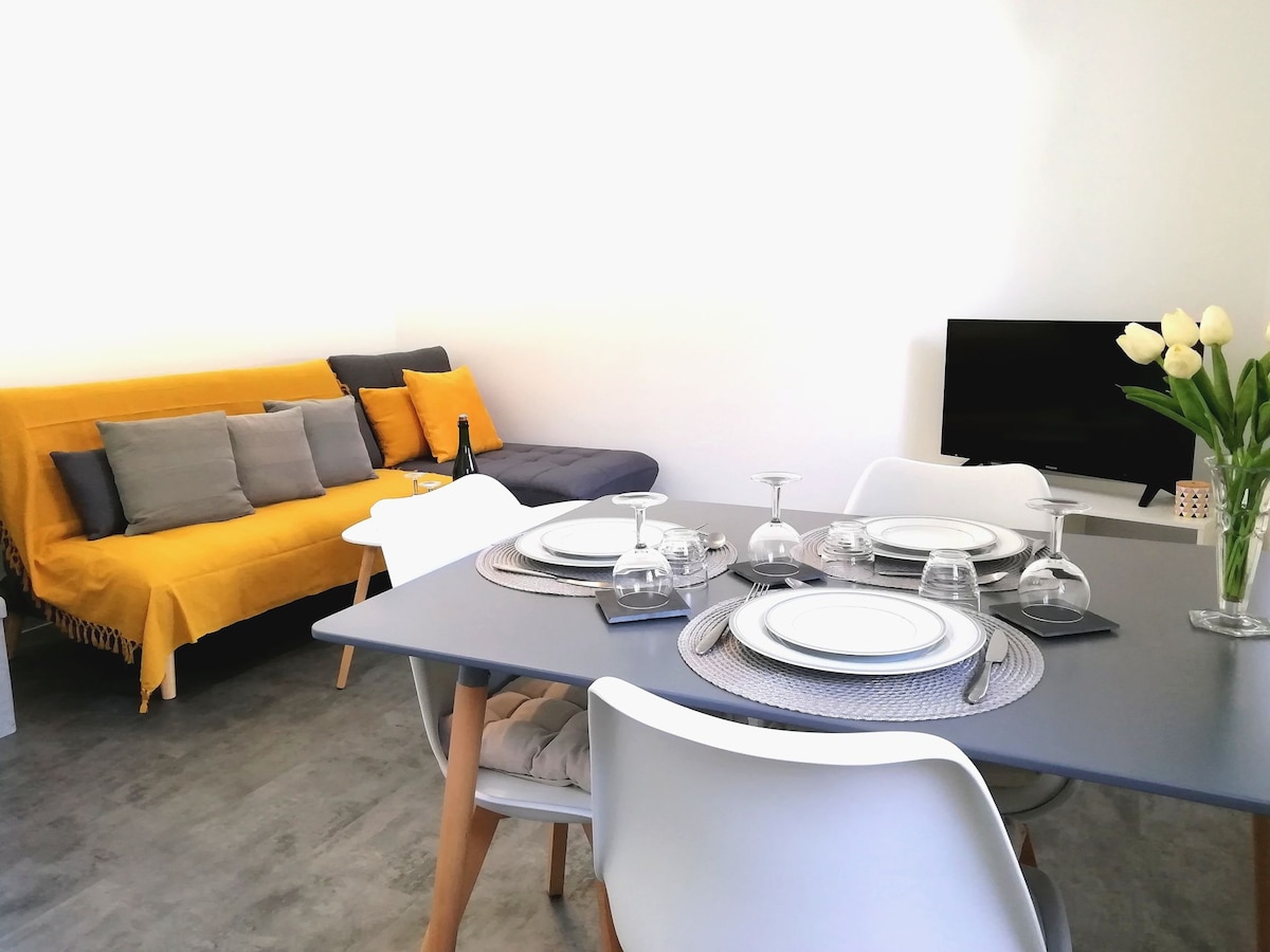 A dining area features a modern table set for four, with plates, glasses, and cutlery neatly arranged. A cozy sofa adorned with yellow and gray cushions is visible in the background, alongside a television, providing a welcoming space for relaxation and dining.