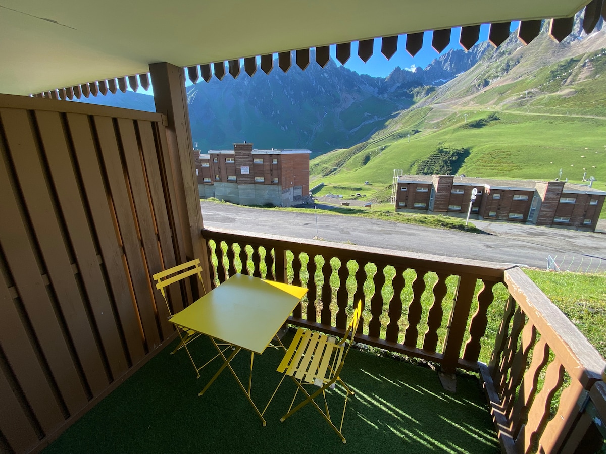 A balcony area features a small yellow table and two matching chairs, overlooking green hills and distant mountain peaks. The surrounding wooden railing adds character, while the scenery includes a glimpse of nearby buildings, contributing to the location's alpine ambiance.
