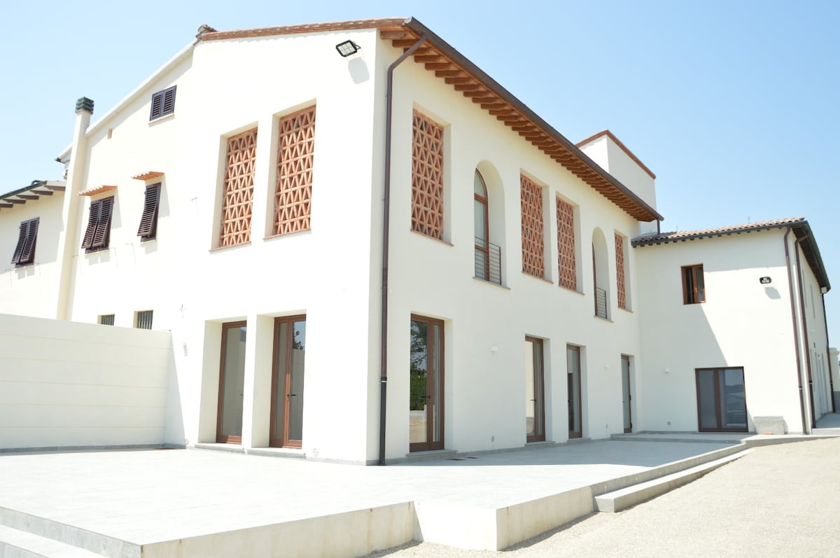 The exterior of the building features a modern design with a white facade accented by patterned brickwork and wooden details. Large windows allow for natural light, while a spacious paved area is visible in front, providing access to various entries.