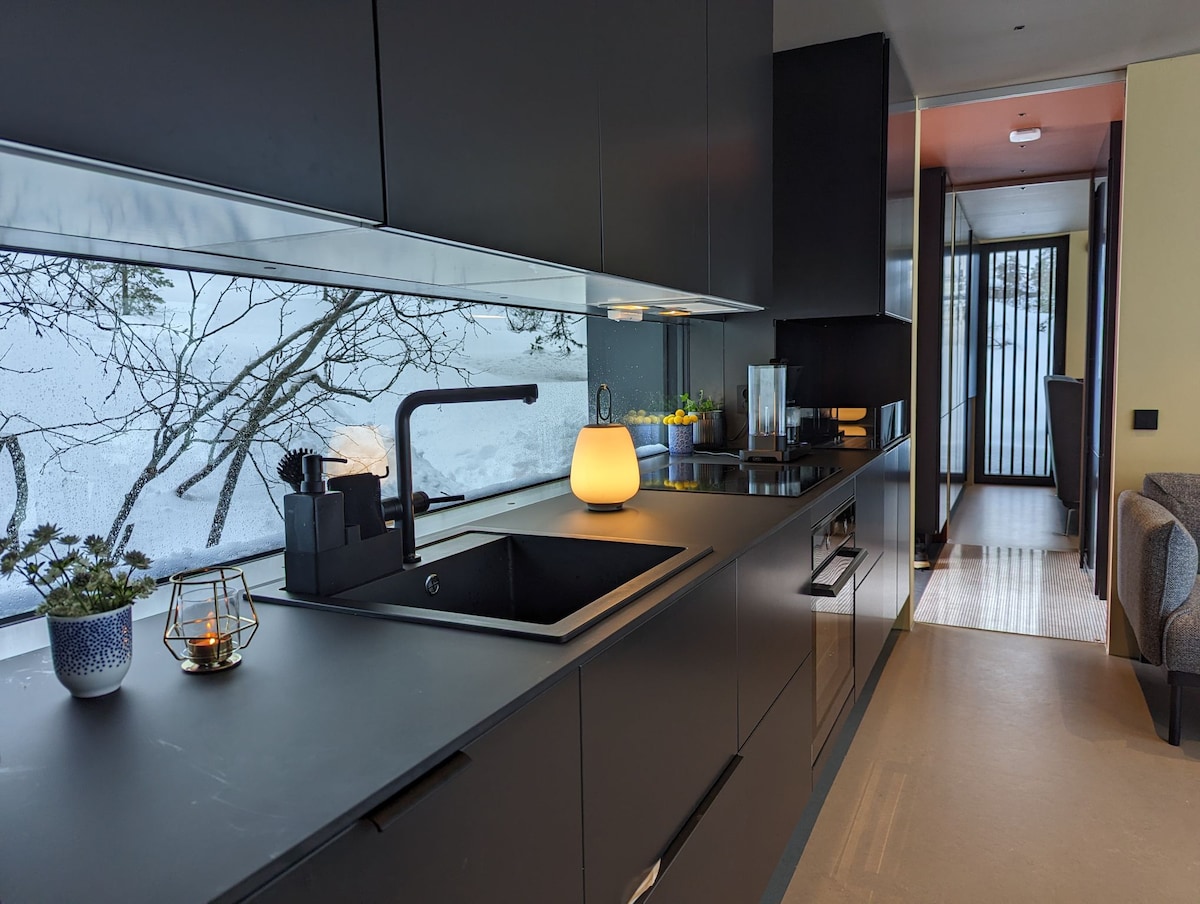 A modern kitchen is presented with sleek black cabinetry and polished surfaces. A window offers views of tree branches, while light from a round lamp illuminates the workspace. Essential appliances, including a stovetop and a coffee maker, are incorporated for convenience.