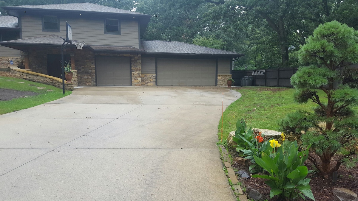 The exterior shows a two-story home with stone accents and a gray facade. A driveway leads to a double garage, and a basketball hoop is visible. Lush greenery surrounds the entrance, featuring a flower bed with colorful blooms and neatly trimmed shrubs.