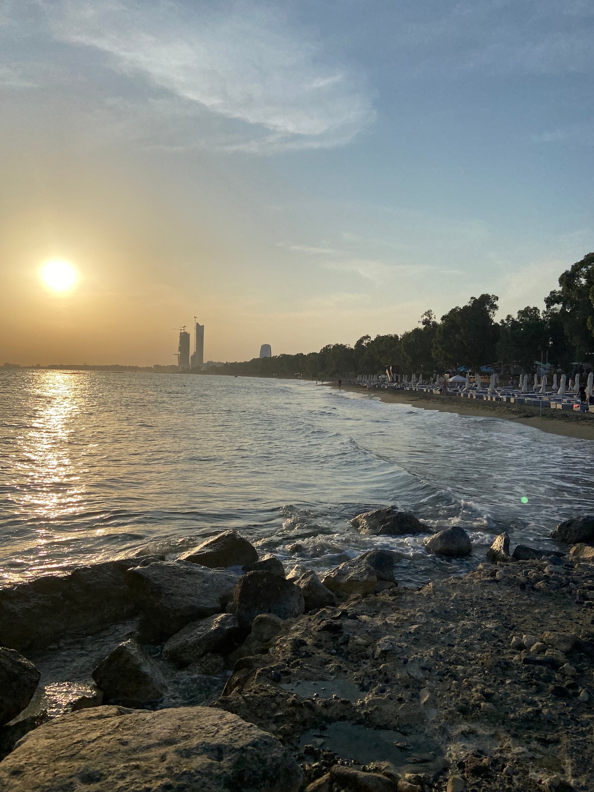 A serene beach scene with gentle waves lapping against rocky shorelines. The setting sun casts a warm glow on the water, reflecting its light toward the horizon. Palm trees and beach umbrellas are visible in the background, creating a tranquil coastal atmosphere.