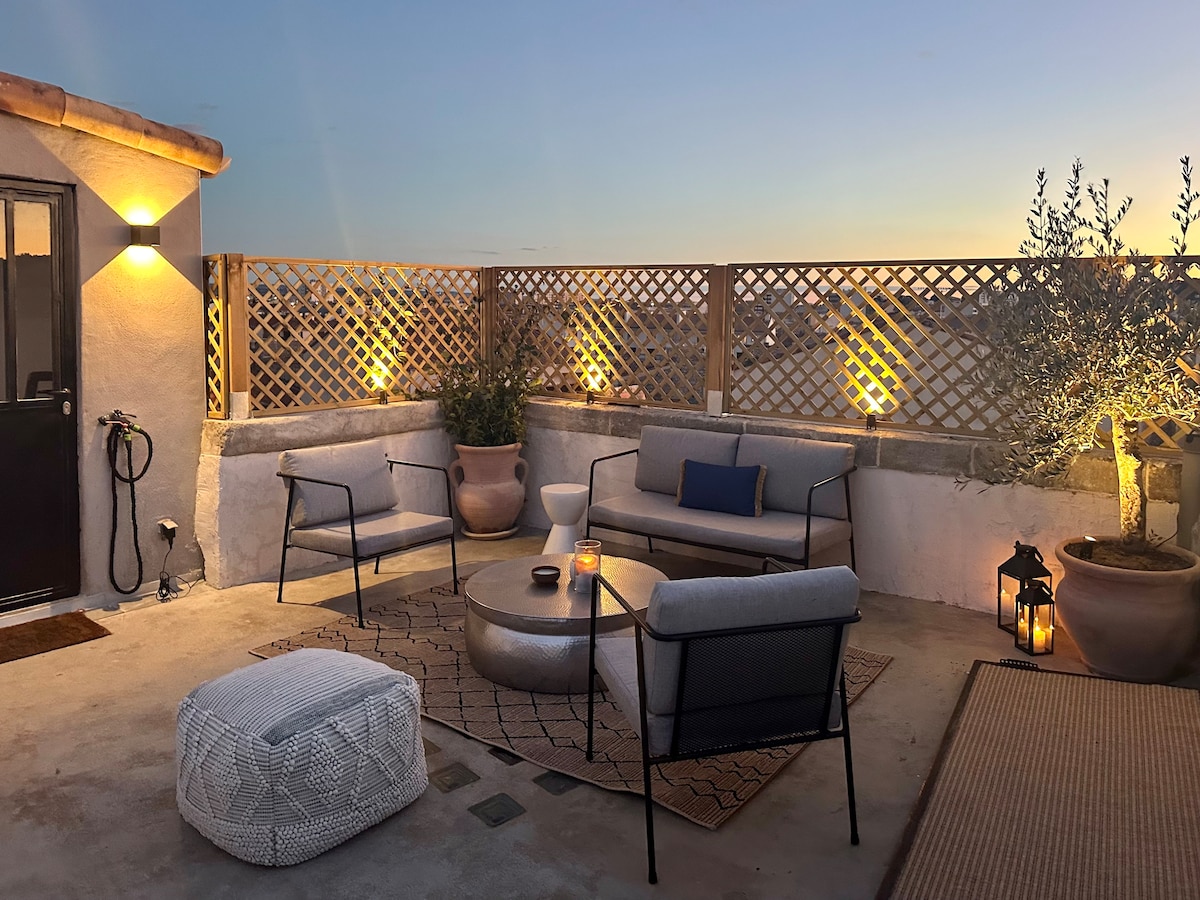 A spacious terrace features comfortable seating arrangements, including two armchairs and a low table, set against a backdrop of a trellis. Soft lighting from wall sconce illuminates the area, complemented by potted greenery and a lantern, creating a relaxed outdoor ambiance.