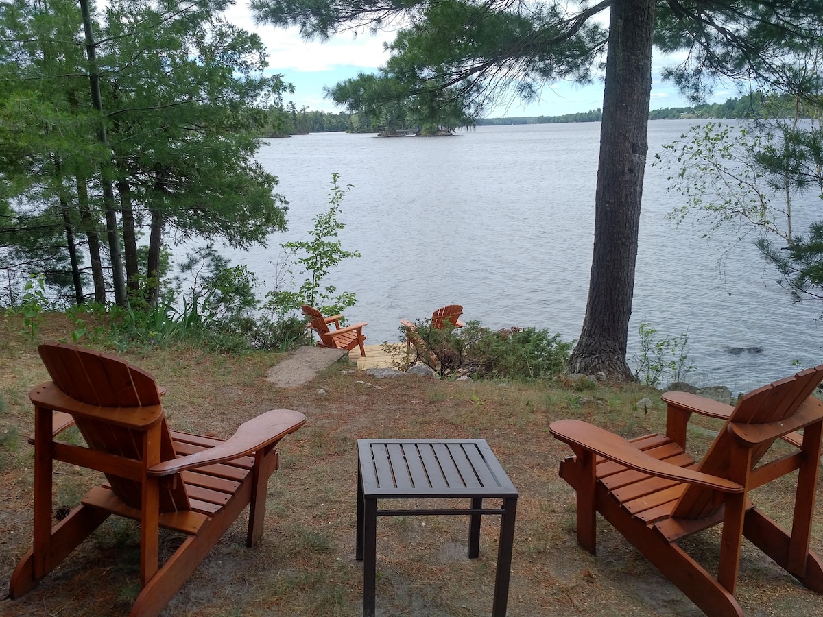 Two wooden adirondack chairs are positioned on a grassy area overlooking the water. A small table is placed between them. Surrounding trees provide shade, while a sandy section of the beach can be seen in the distance, contributing to the natural setting.