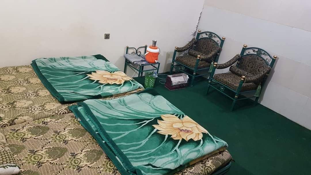 A cozy sleeping area is furnished with two beds covered by green blankets featuring large floral designs. Two armchairs with intricate patterns are situated near a small table, offering a comfortable seating arrangement. The room is carpeted in dark green.