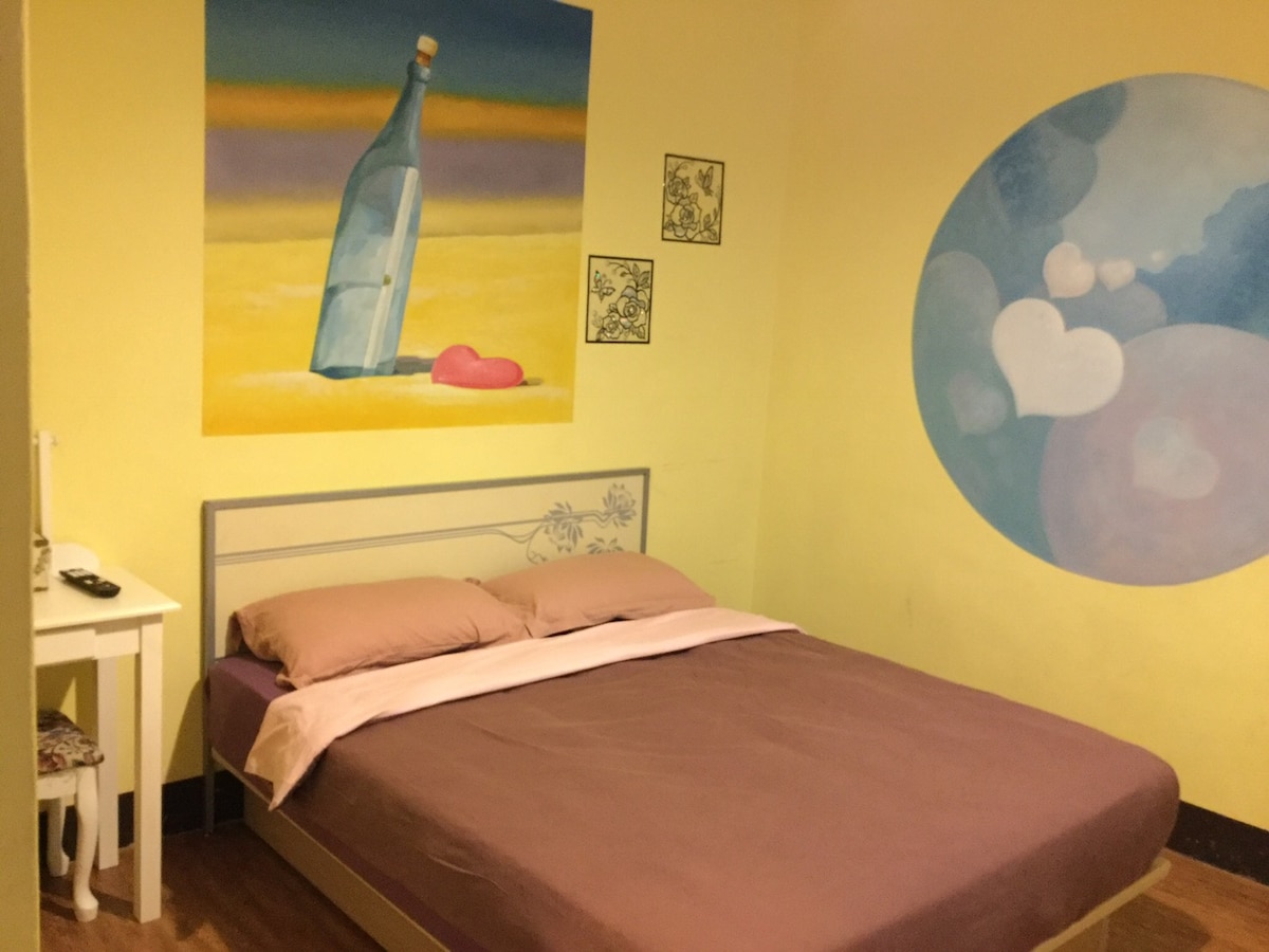 A cozy room features a spacious bed with a soft pink blanket. Vibrant yellow walls are adorned with a large mural depicting a bottle and heart shapes, contributing to a bright and cheerful ambiance. A small bedside table complements the decor.
