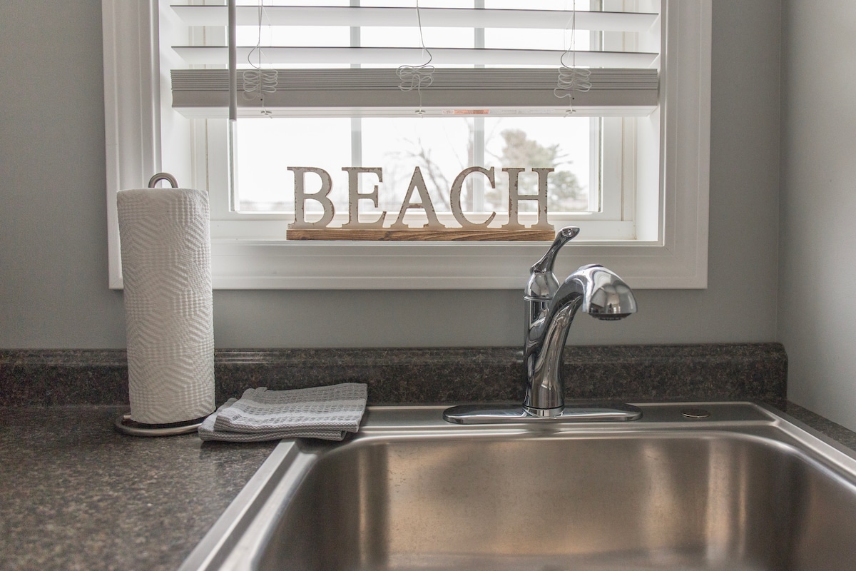 A kitchen sink is positioned beneath a window adorned with white blinds. The countertop is dark and features a roll of paper towels and a gray dish towel. A wooden sign spelling 'BEACH' is displayed on the sill, offering a subtle coastal theme.