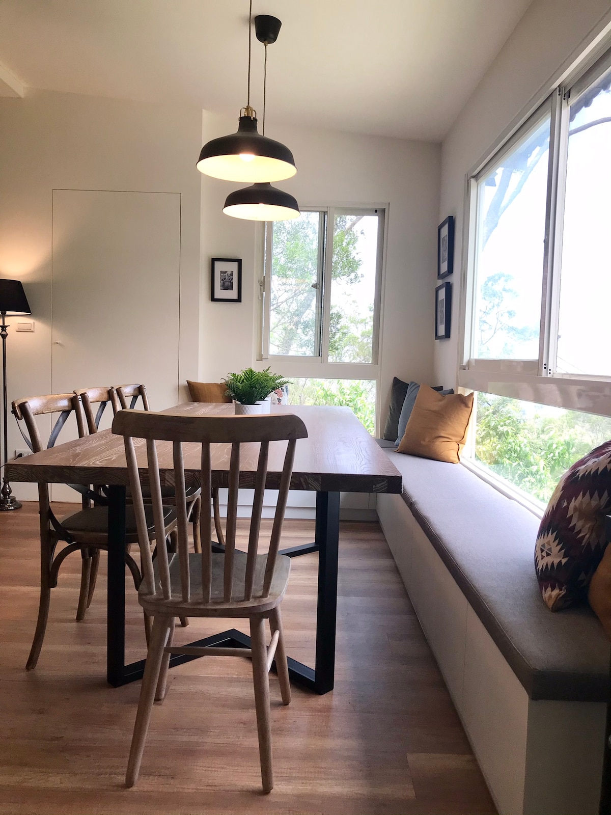 A dining area features a wooden table with matching chairs and a cozy built-in bench adorned with cushions. Large windows frame the space, allowing natural light to brighten the room and providing views of the surrounding greenery.
