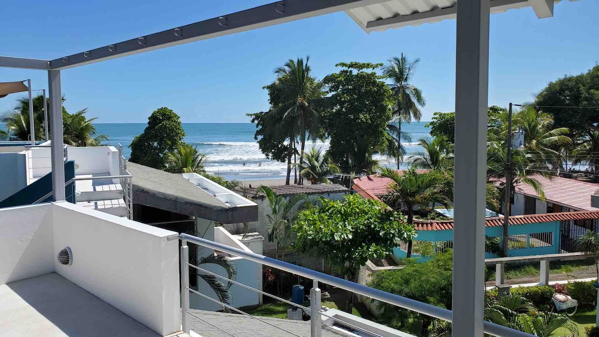 A covered upper patio offers a panoramic view of the ocean and surrounding palm trees. The gently rolling waves are visible in the distance, and lush greenery surrounds the area, highlighting the tranquil beachfront environment.