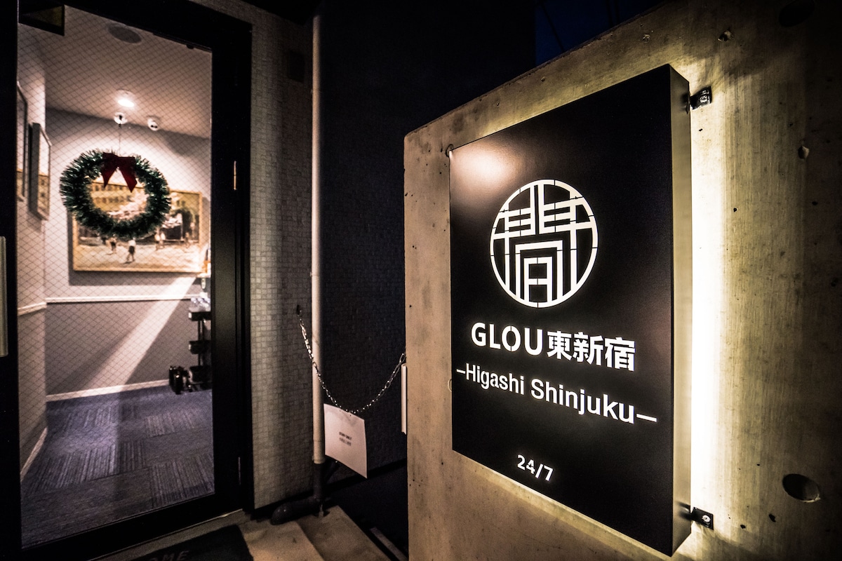 The entrance to the lodging is marked by a modern, illuminated sign featuring the name 'GLOU 東新宿.' A welcoming atmosphere is suggested by a festive wreath positioned beside the door, leading into a well-lit lobby area.