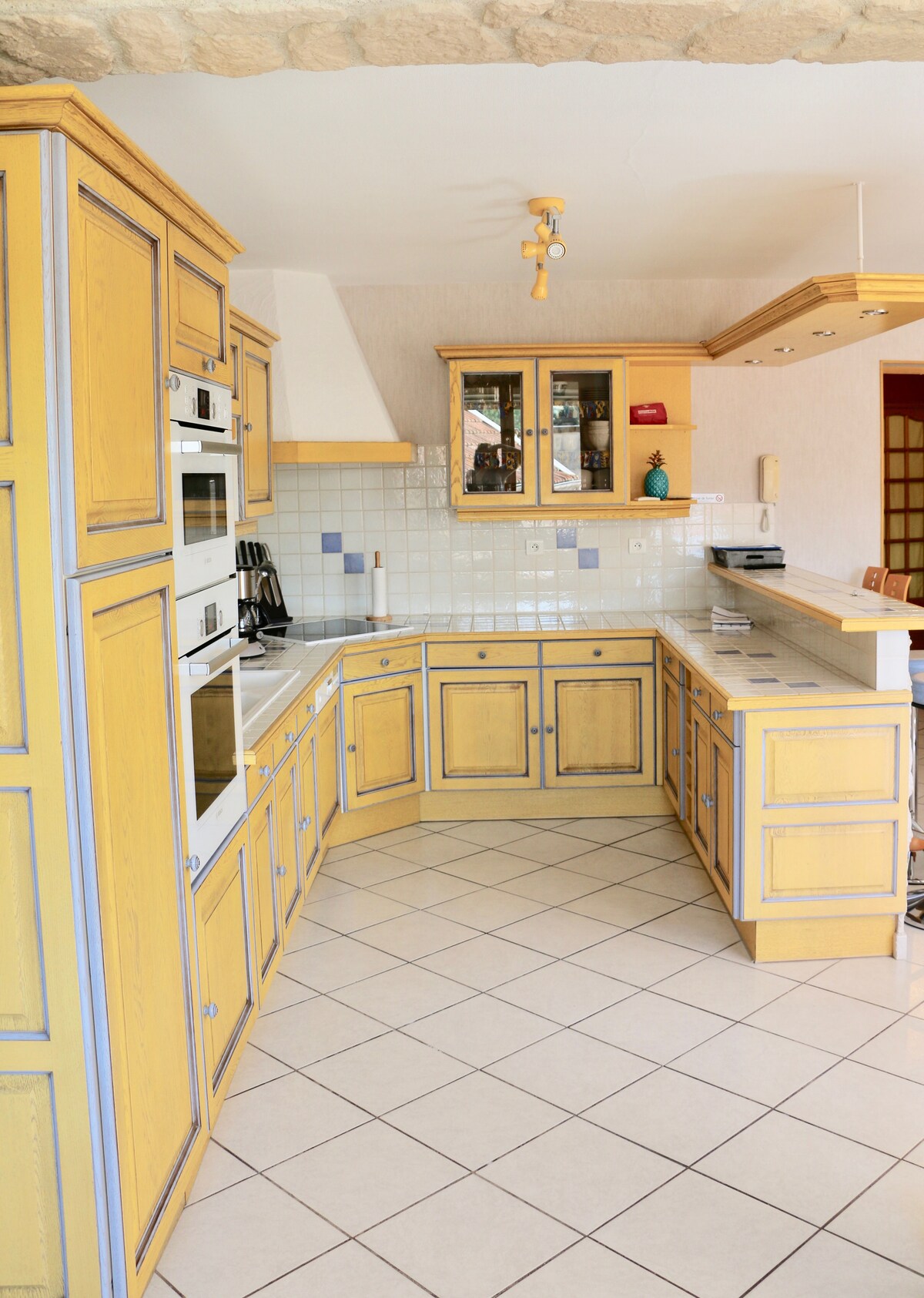 An equipped open kitchen features yellow cabinetry with blue accents. Modern appliances, including ovens and a stove, are integrated into the design. The tiled floor reflects natural light, contributing to the spacious atmosphere. Clear wall tiles complement the cabinetry, enhancing practicality.