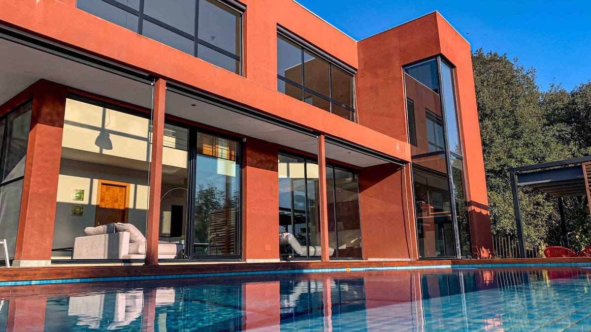A modern structure with large glass windows offers a clear view of the interior spaces. The red exterior contrasts with the blue of the salt-water pool, reflecting the surrounding greenery. Sunlight brightens the area, enhancing the spacious layout of the design.