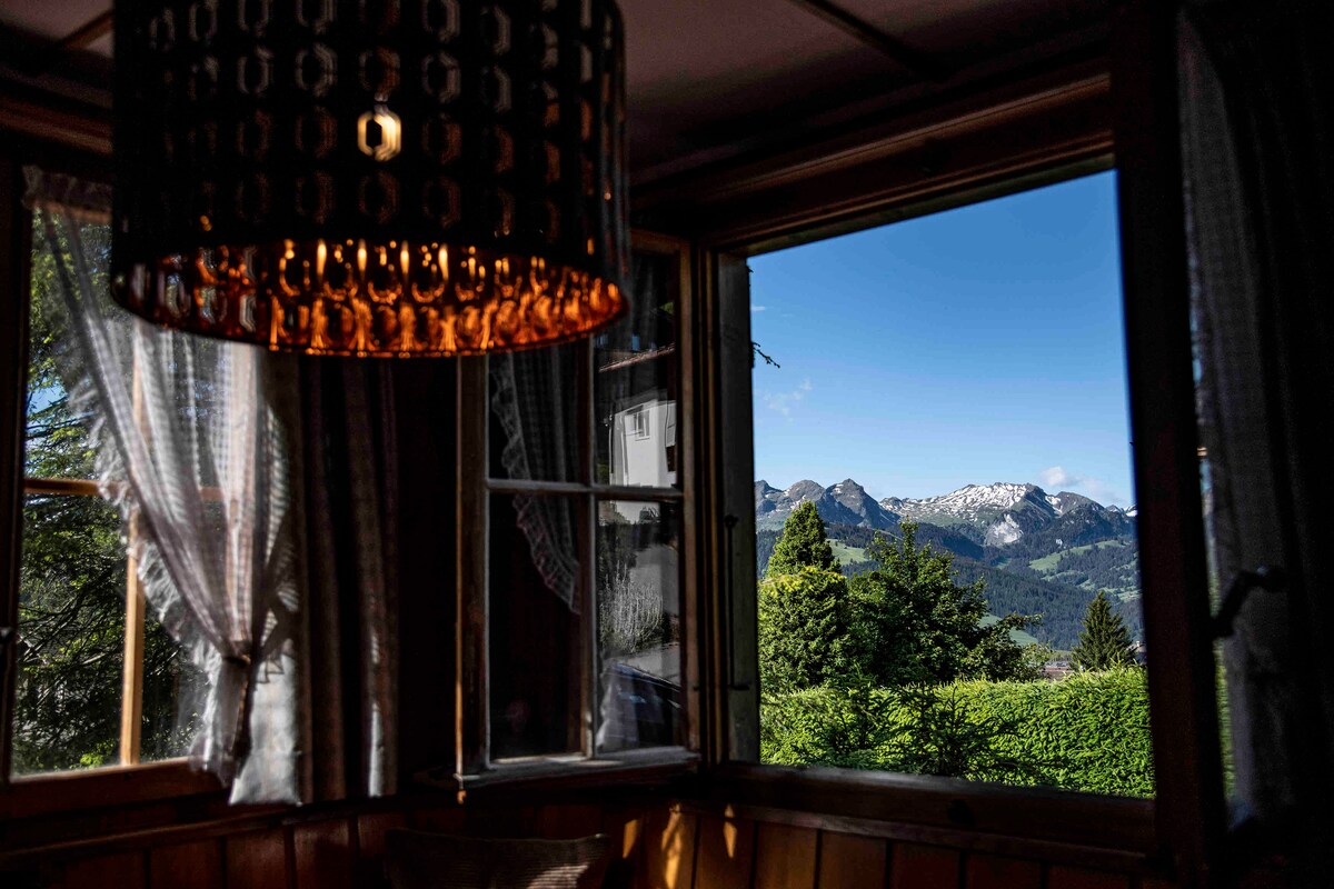 A scenic view is framed by two large windows, offering a glimpse of majestic mountains and lush greenery. Natural light enhances the interior, where a decorative chandelier adds warmth to the space. Sheer curtains gently diffuse the sunlight, creating a serene ambiance.