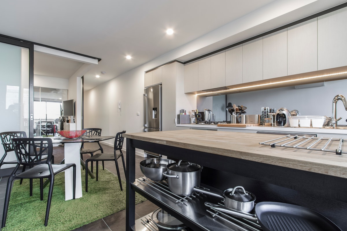 A modern kitchen is displayed, featuring sleek cabinetry and stainless-steel appliances. A large island with a wooden countertop is positioned in the foreground, showcasing cookware and utensils. A dining area is included with four black chairs surrounding a white table, enhancing the spacious layout.