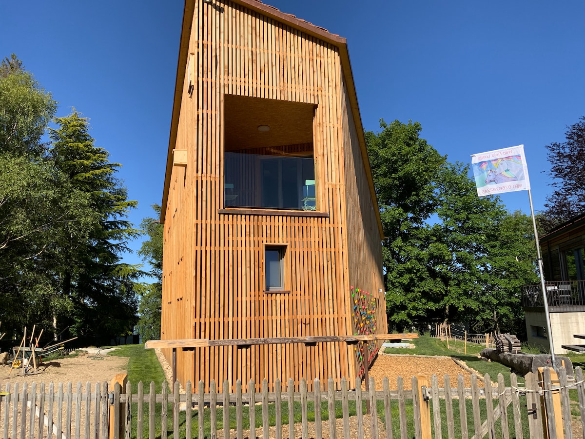 The wooden building features a unique sloped design with vertical wooden slats, allowing natural light to filter through. A large window and a smaller window provide views of the surrounding greenery. A climbing wall is visible on one side, enhancing outdoor recreational opportunities.
