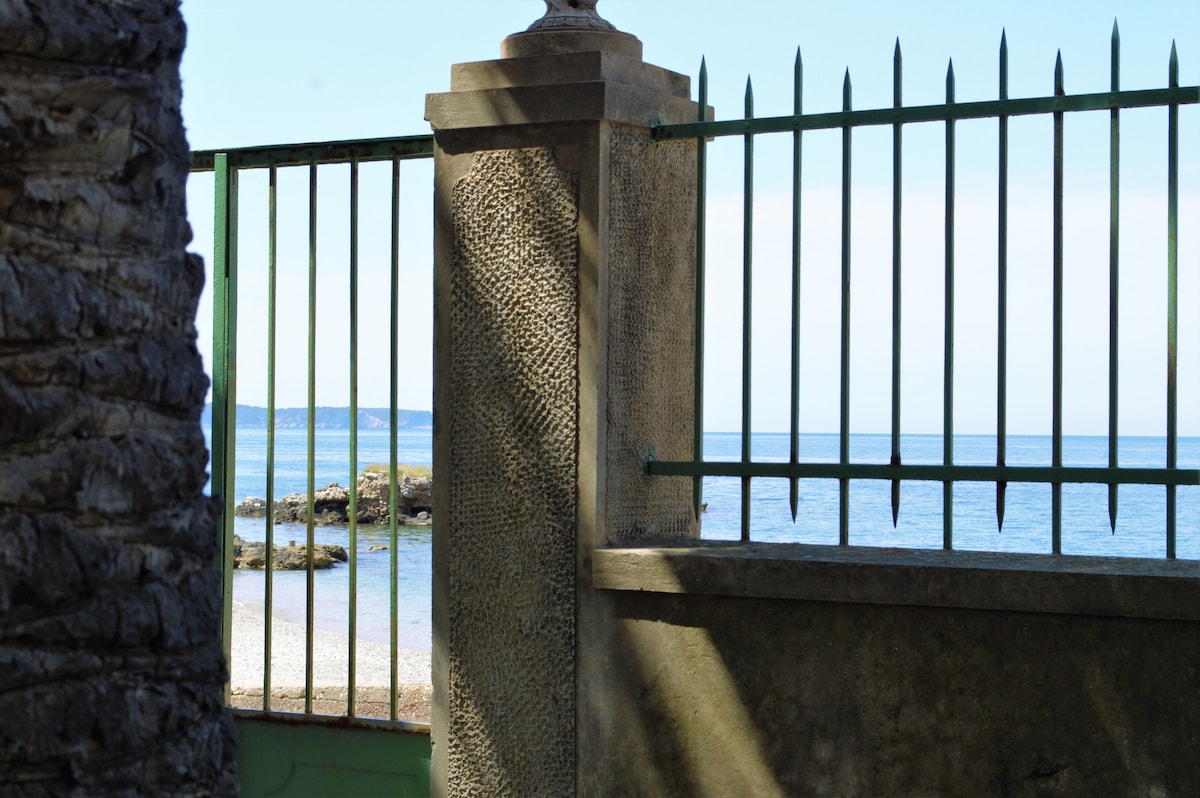 A green metal gate is framed by a textured stone column, leading to a serene view of the calm sea. The beach is visible through the gate, where gentle waves lap against the shore under a clear blue sky.