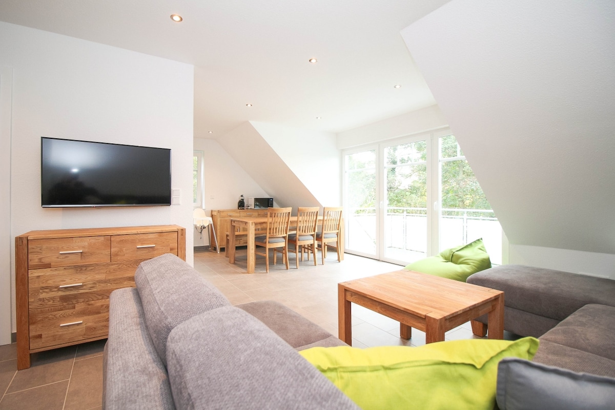 A light-filled living area features a large, comfortable sofa with bright green accent pillows. A wooden dining table with matching chairs is visible, alongside a flat-screen television mounted on the wall. Large windows allow natural light to illuminate the space.