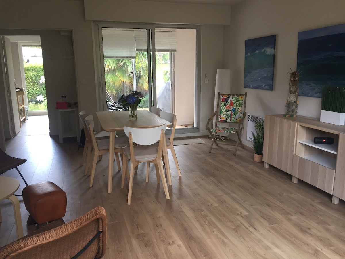 A bright living space features a dining table surrounded by six light wooden chairs. Large sliding doors provide access to a terrace, while two artworks depicting ocean waves are displayed on the wall. A sideboard and comfortable seating are arranged to enhance the openness.