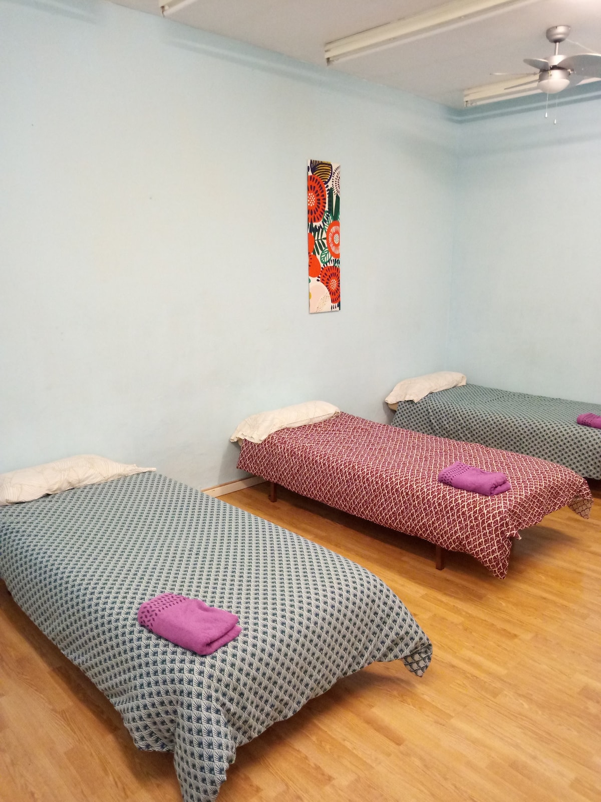 A spacious room contains three beds with vibrant patterned coverings. Each bed is arranged with a towel rolled neatly at the foot. A light blue wall adds brightness to the space, complemented by wooden flooring and a colorful piece of wall art.