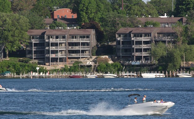 A view of the condo complex is presented across the water, featuring multilevel buildings with balconies. A boat is seen gliding smoothly over the lake's surface, surrounded by lush greenery and additional boats docked nearby. The natural setting creates a calm atmosphere.