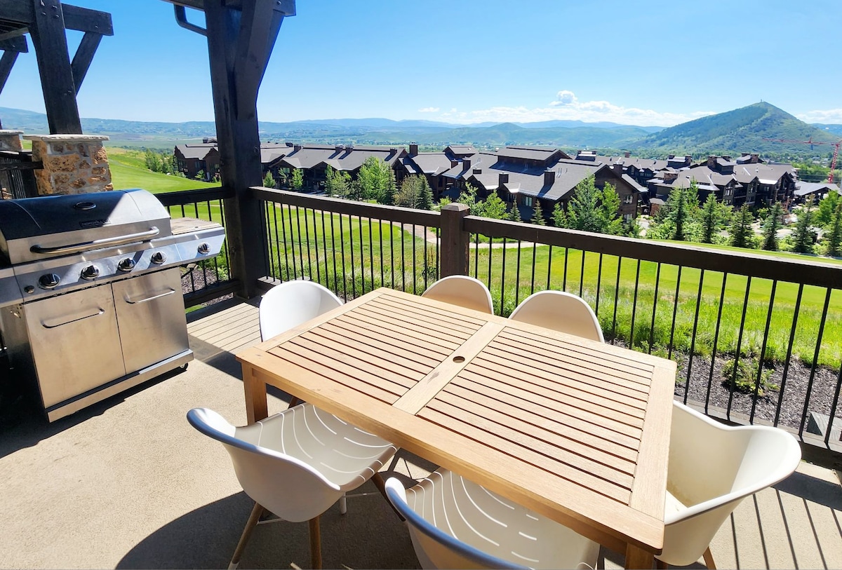 An outdoor terrace showcases a wooden dining table surrounded by four white chairs. A stainless steel grill is positioned nearby, with expansive views of the rolling hills and mountain backdrop visible beyond the railing, creating a serene outdoor dining space.