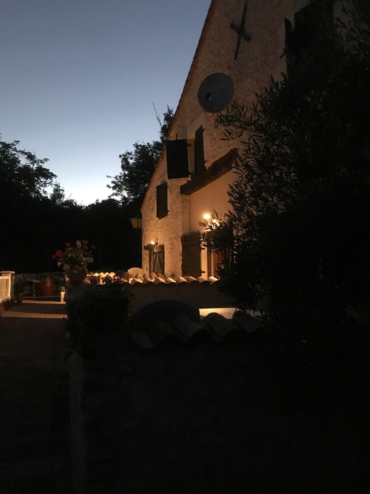 The stone exterior of the house is softly illuminated as dusk sets in, highlighting the rustic architecture. Potted plants can be seen along the terrace, and a gentle glow emanates from window fixtures, creating a serene atmosphere in the surrounding garden.
