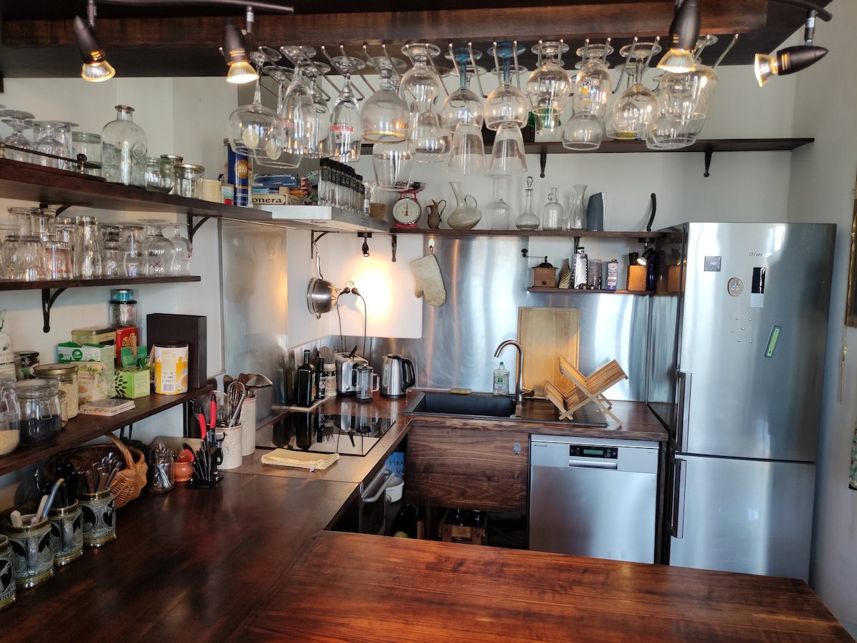 A well-equipped kitchen features dark wooden countertops and open shelving holding various glassware. Cookware and appliances are neatly arranged, including a stainless steel refrigerator and a dishwasher. Above, glassware is suspended from a hanging rack, enhancing the functional and organized layout.