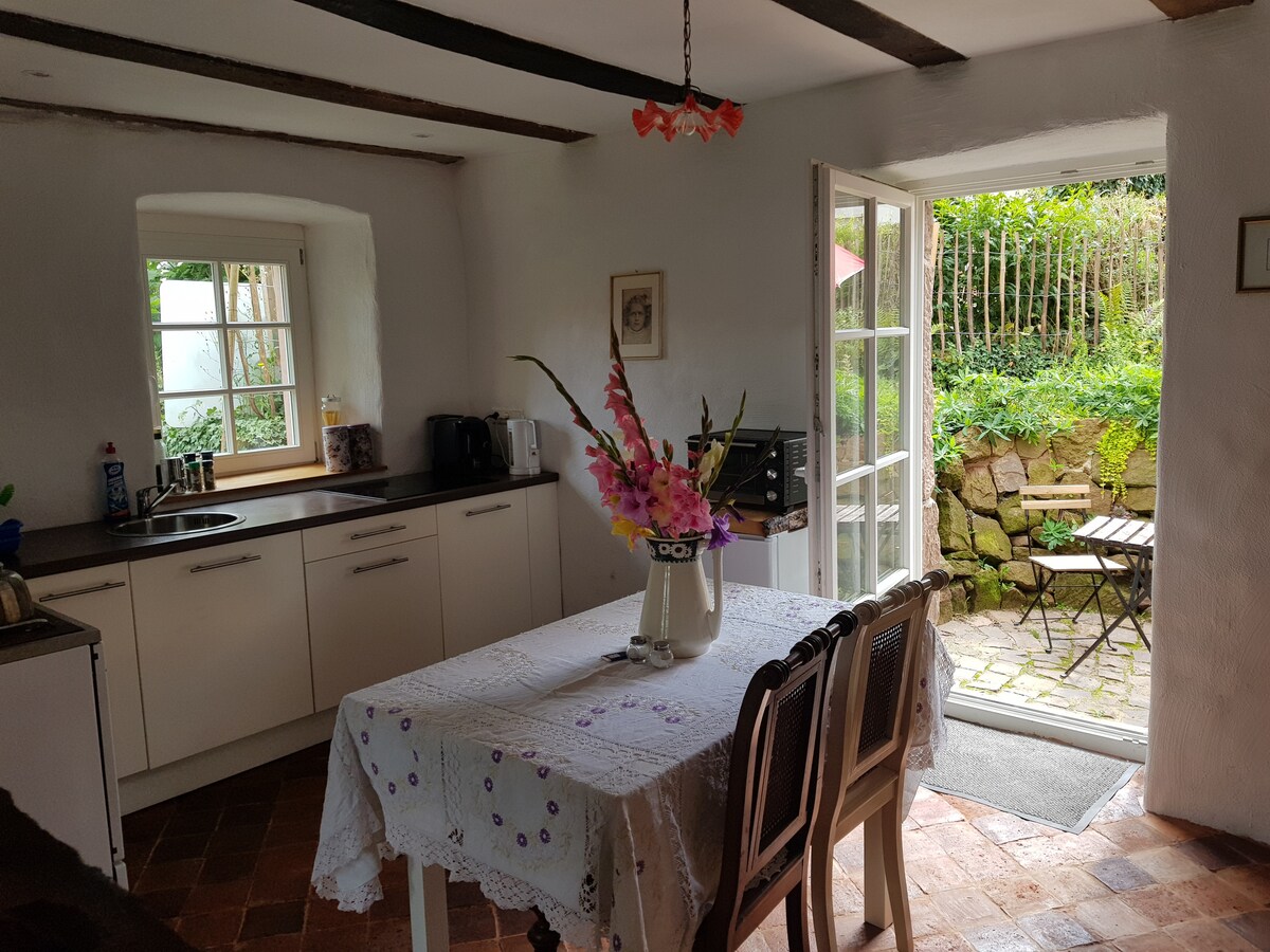 A bright and airy kitchen area features white walls and wooden beams overhead. A dining table, adorned with a floral centerpiece, is surrounded by four chairs. Glass doors open to a small patio, revealing a stone garden with outdoor seating.