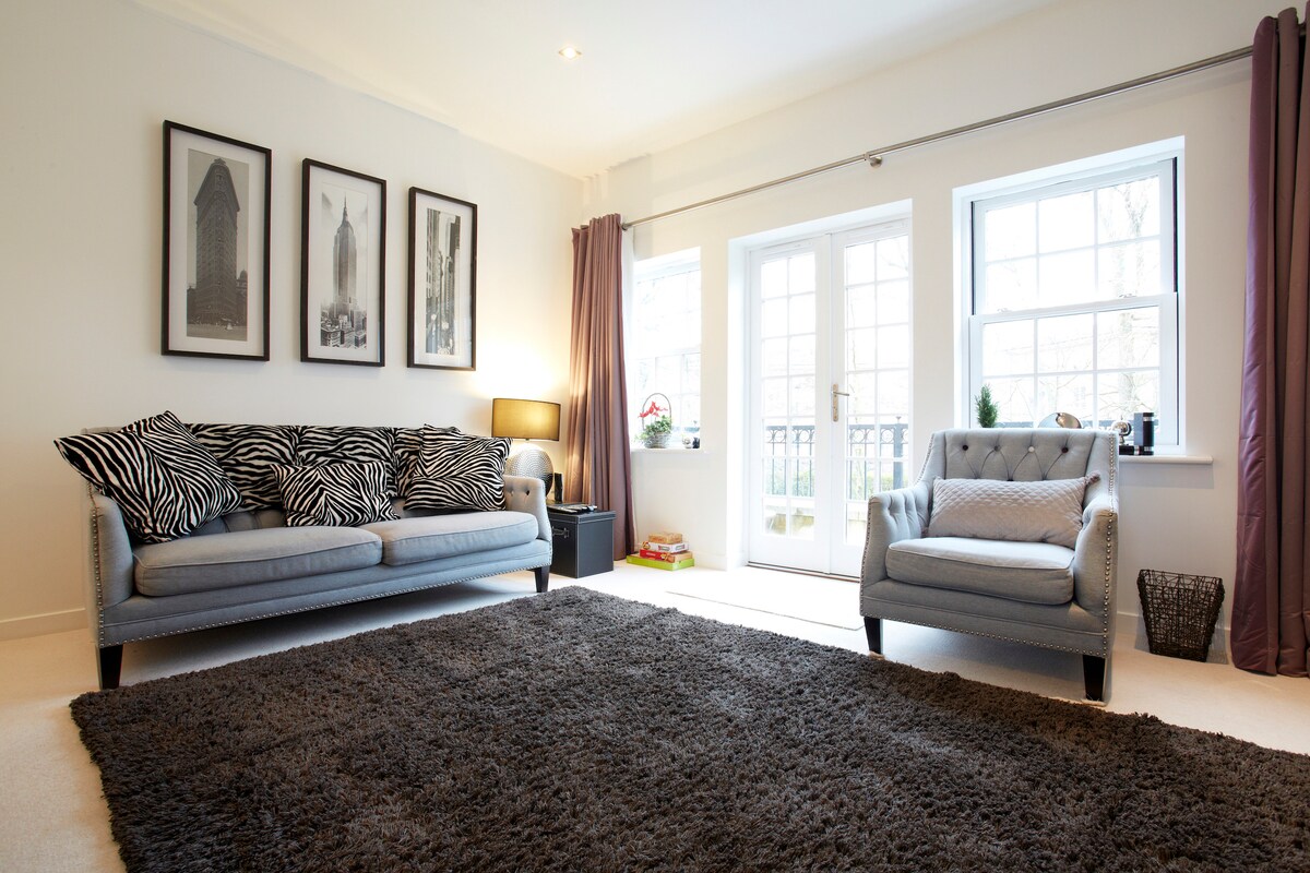 A bright living area is furnished with a zebra-patterned sofa and a single upholstered chair. Natural light filters in through double doors, leading to a balcony. Framed art pieces adorn the wall, complemented by a soft, textured rug spanning the floor.