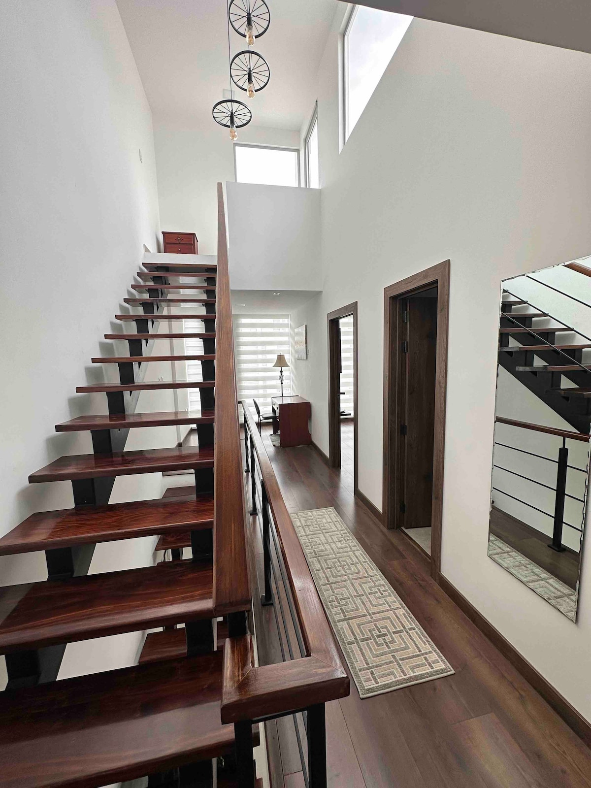 A well-lit entrance features an elegantly designed staircase with wooden steps leading to the upper level. A minimalist rug lines the smooth floor, while a mirror reflects the natural light from high windows. Additional doors provide access to adjacent rooms.