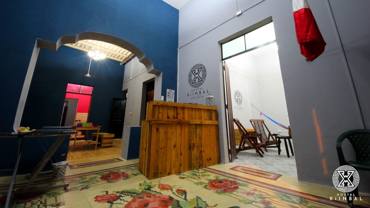 A communal area is presented with a wooden reception desk, rustic seating, and colorful tiled flooring. The walls are painted in calming shades, featuring the hostal's logo. Natural light streams through an entrance, while a traditional Mexican rug adds warmth to the space.