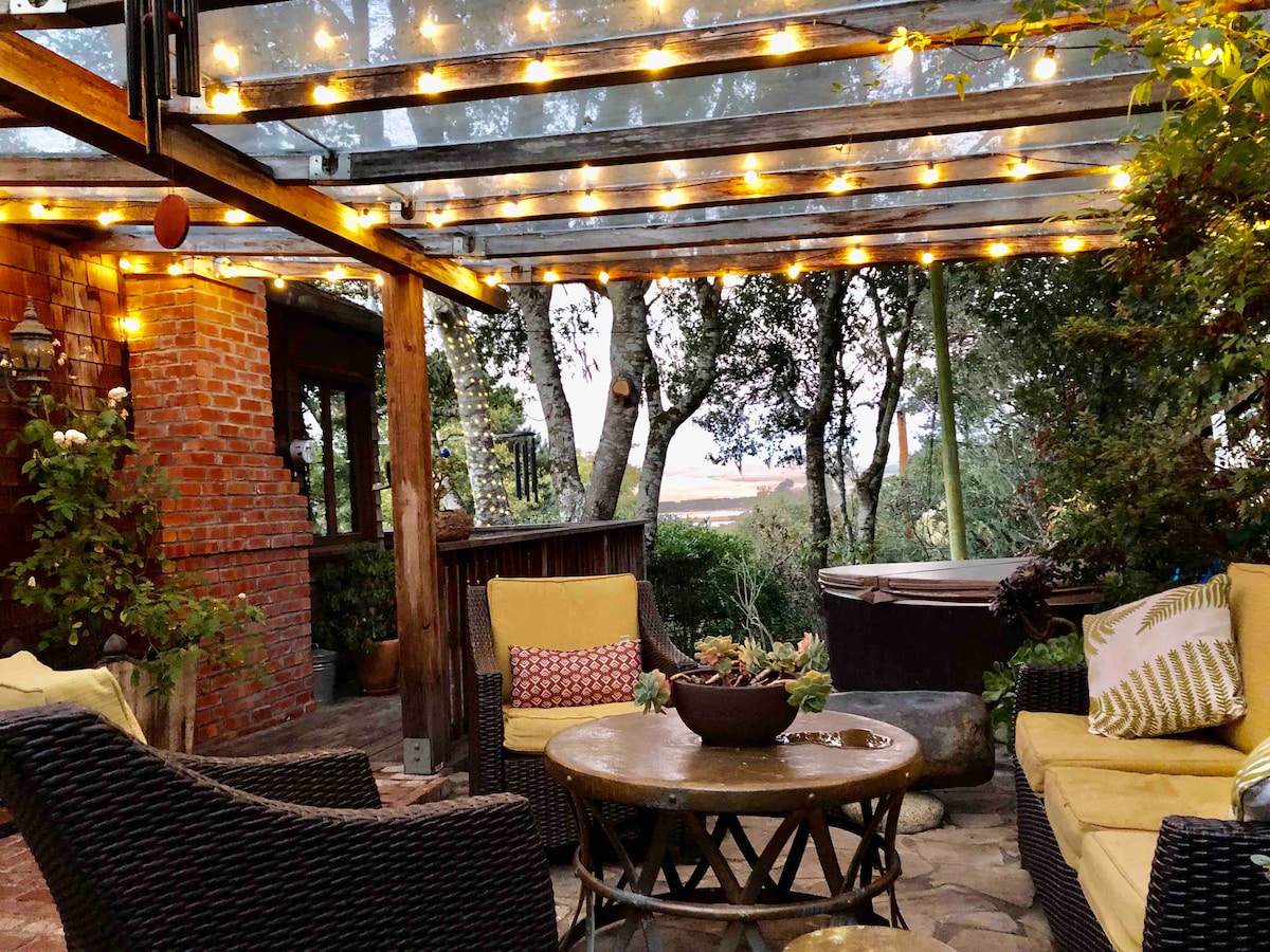 An inviting outdoor lounge area is seen under a covered pergola adorned with soft twinkle lights. Comfortable seating is arranged around a circular table, complemented by colorful cushions. A hot tub and surrounding greenery add to the ambiance of relaxation and comfort.