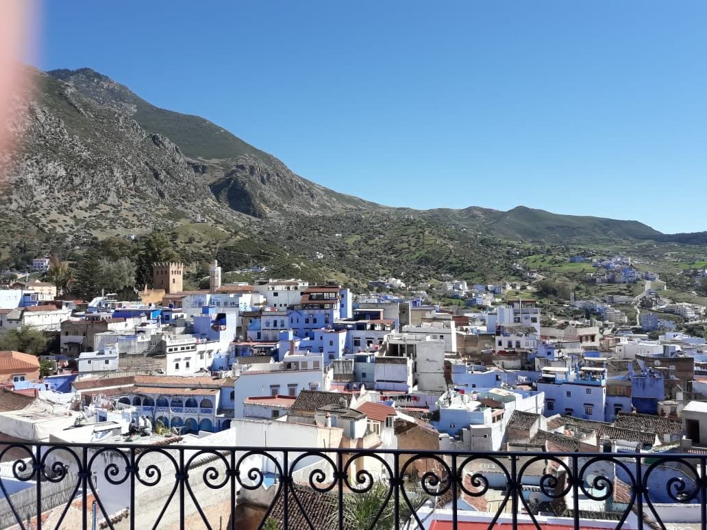 A panoramic view captures the vibrant buildings of Chefchaouen, with their characteristic blue hues. The backdrop features a mountainous landscape under a clear blue sky. A decorative iron railing frames the image, accentuating the scenic vista.