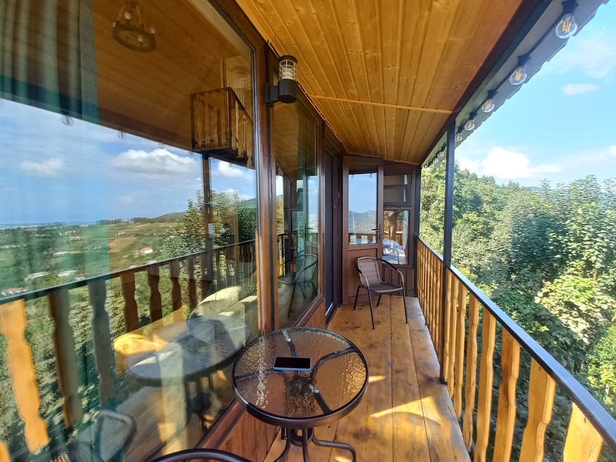 A wooden balcony is framed by large glass windows, offering panoramic views of the surrounding landscape. A round glass-top table and two wicker chairs are arranged on the floor, and soft lighting fixtures hang from the wooden ceiling.