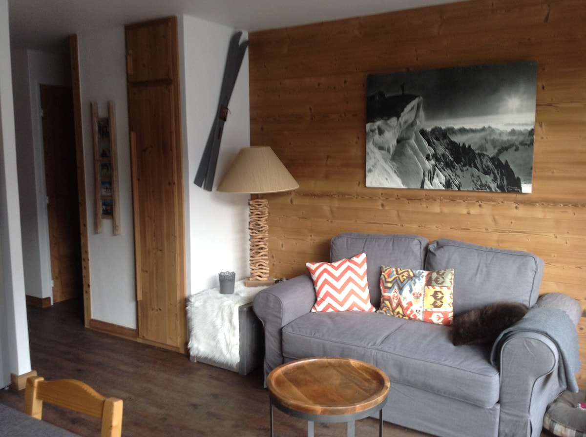 A cozy living area features a gray sofa adorned with colorful cushions, alongside a wooden coffee table. A rustic lamp stands beside the sofa, and a wall-mounted ski decor adds to the charm. Natural light illuminates the wood-paneled wall, creating a warm atmosphere.