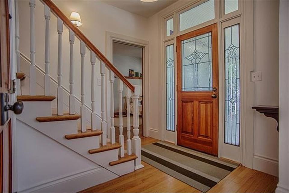 An inviting foyer features a wooden front door with decorative glass panels, flanked by two tall windows. A striped rug lies beneath a staircase, which is adorned with smooth wooden railings, leading up to the second floor.