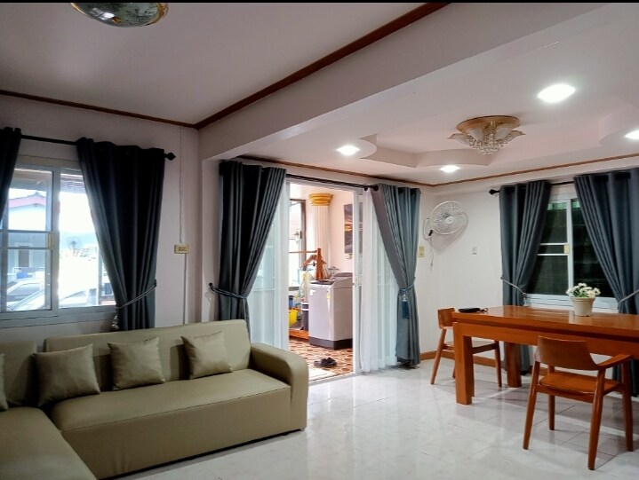 A modern living area is present, featuring a comfortable sofa and a dining table with wooden chairs. Large windows allow natural light to fill the space, while grey curtains frame the windows. An open doorway leads to a bright kitchen area.
