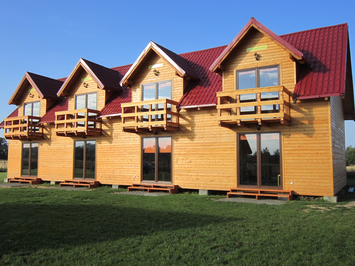 A row of wooden cabins is displayed, each featuring a pitched roof with bright red shingles. Each cabin includes a balcony that offers views of the surrounding landscape, and large glass doors provide access to the outdoors. The green lawn in front enhances the natural setting.